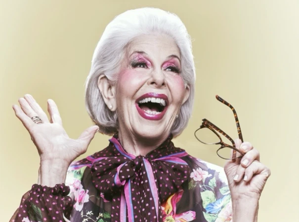 Elderly woman with white hair, makeup, floral top, holding glasses in photo studio smiling with hand raised in front of a yellow backdrop.