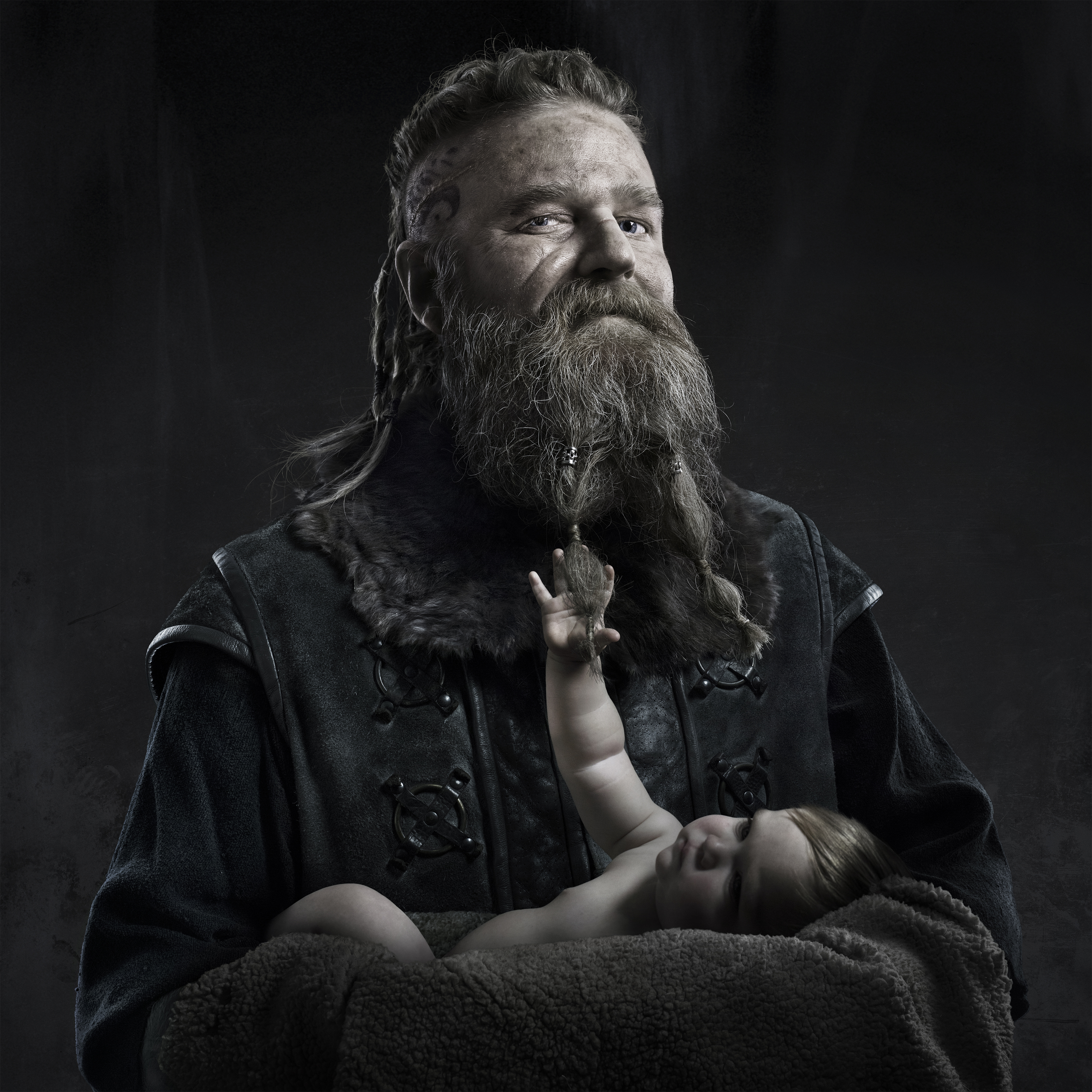 Adult male cradles a sleeping baby in a dark, moody portrait. The man has a long beard and wears a dark, fur-lined coat.