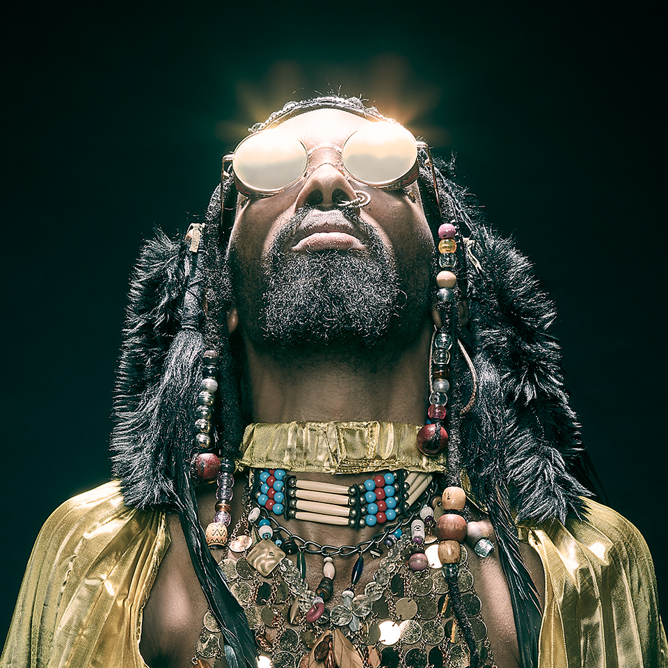 Portrait of black man with beard and sunglasses looking up; wearing gold top, beaded necklaces, and fur trim headpiece on a dark background.