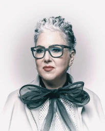 Portrait of an older woman with curly gray hair wearing thick-rimmed glasses and a sheer bow tie by Jeff Sciortino.