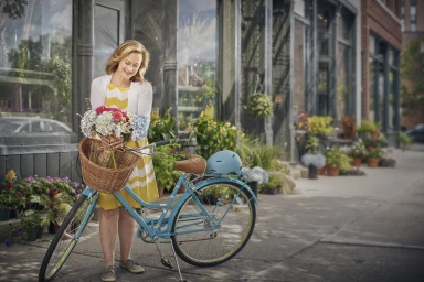 Mature woman holding a bouquet of flowers next to a blue bicycle on a city sidewalk by a storefront.