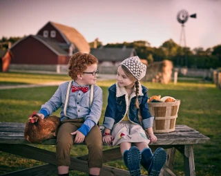 Young boy and girl seated on a wooden picnic table on a farm with a red barn and windmill in the background.