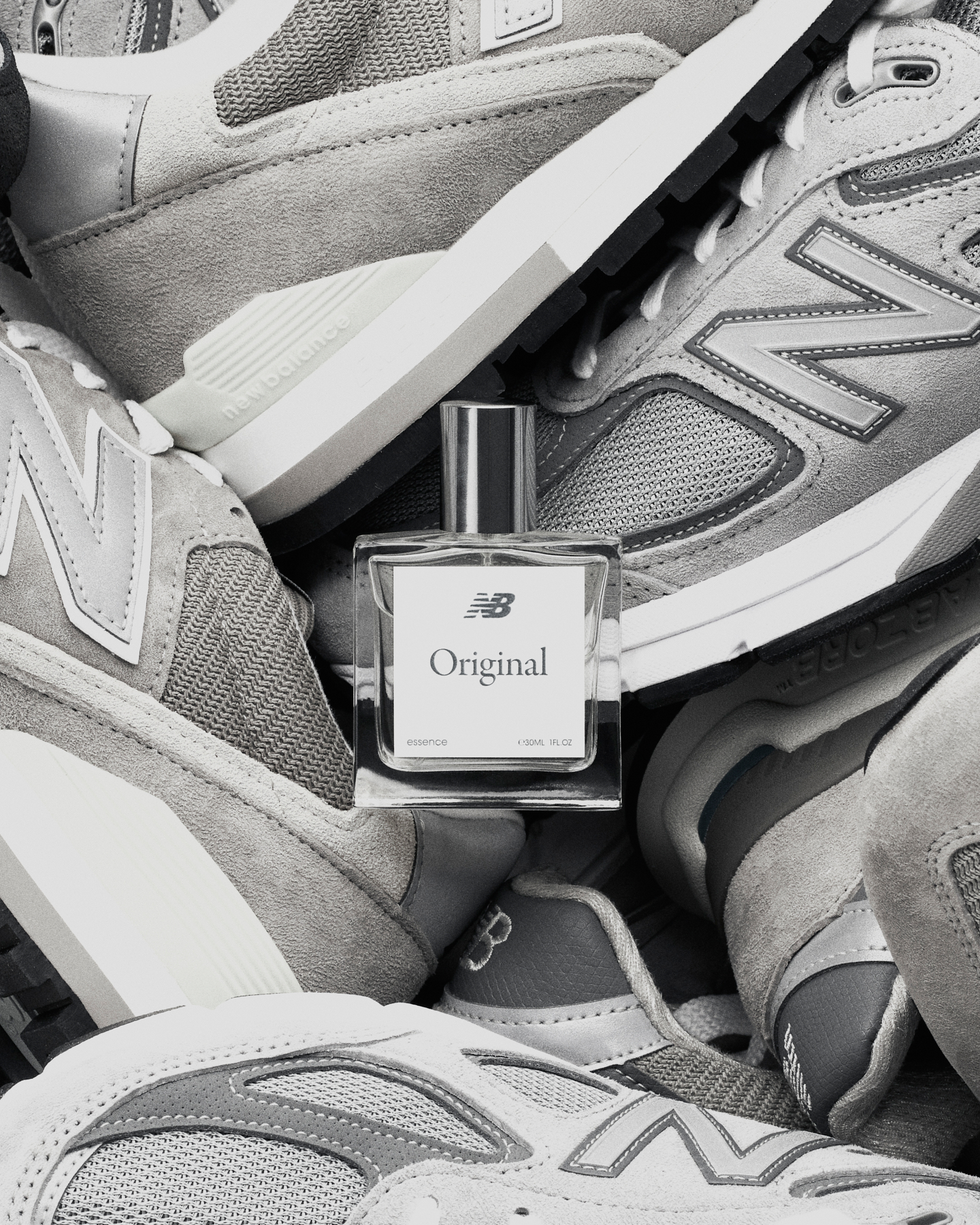 Monochrome product shot featuring a clear glass perfume bottle labeled 'Original' nestled amongst gray New Balance running shoes.