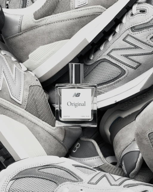 Monochrome product shot featuring a clear glass perfume bottle labeled 'Original' nestled amongst gray New Balance running shoes.