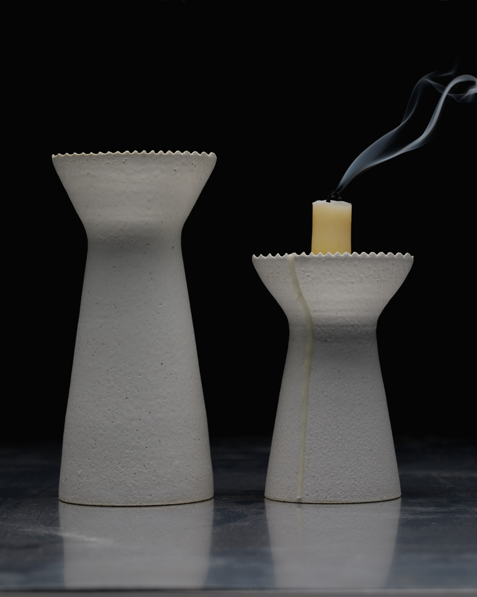 Two textured white candle holders, one holding a lit candle with smoke, are set against a black background, creating a minimal scene.