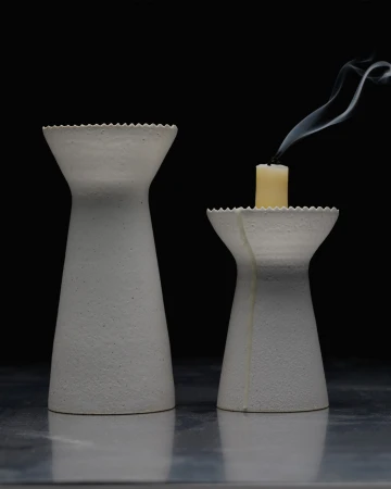 Two textured white candle holders, one holding a lit candle with smoke, are set against a black background, creating a minimal scene.