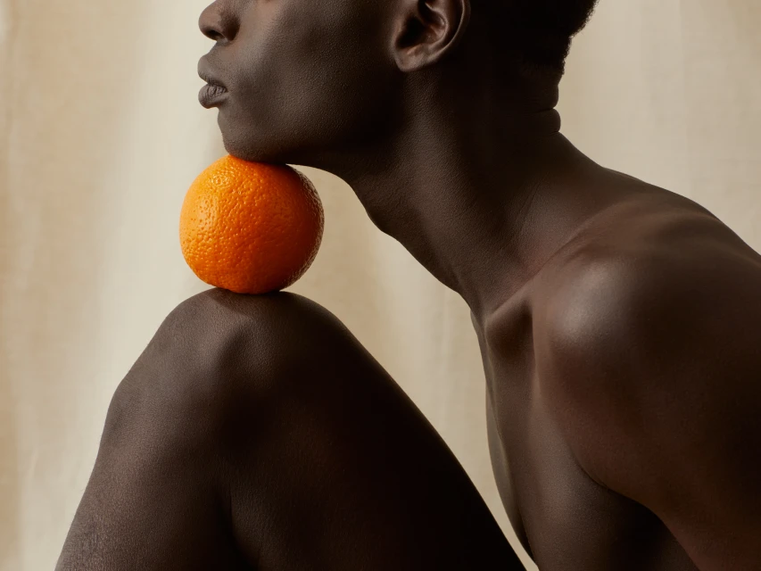 Striking studio portrait of a dark-skinned adult male posing with an orange balancing on his shoulder against a cream backdrop.