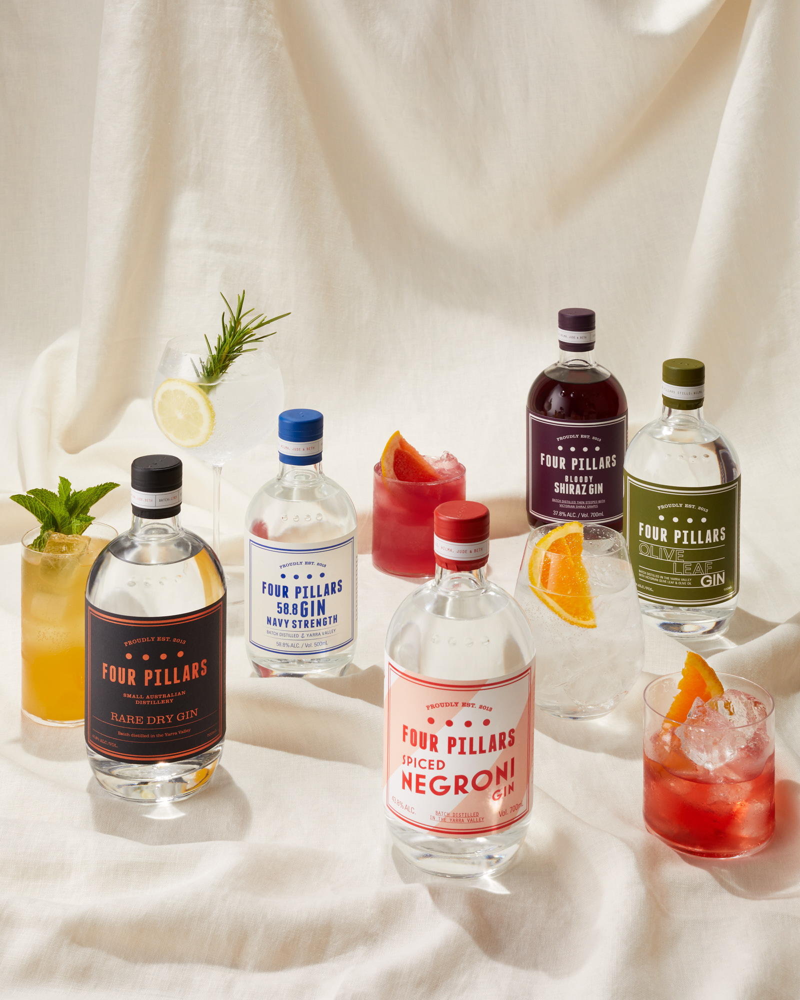Several bottles of Four Pillars gin varieties sit on a white linen surface, with cocktails and citrus garnishes, arranged in a visually appealing composition.