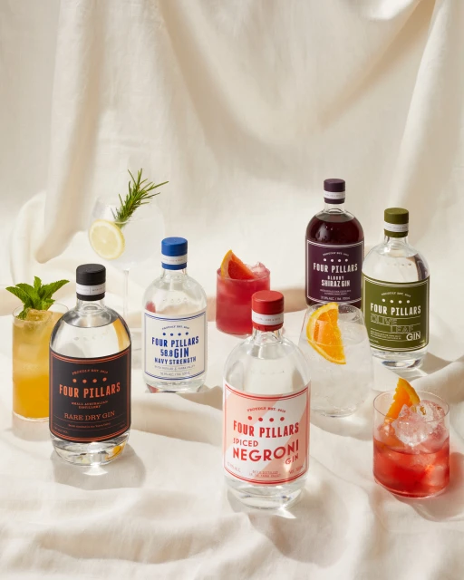 Several bottles of Four Pillars gin varieties sit on a white linen surface, with cocktails and citrus garnishes, arranged in a visually appealing composition.
