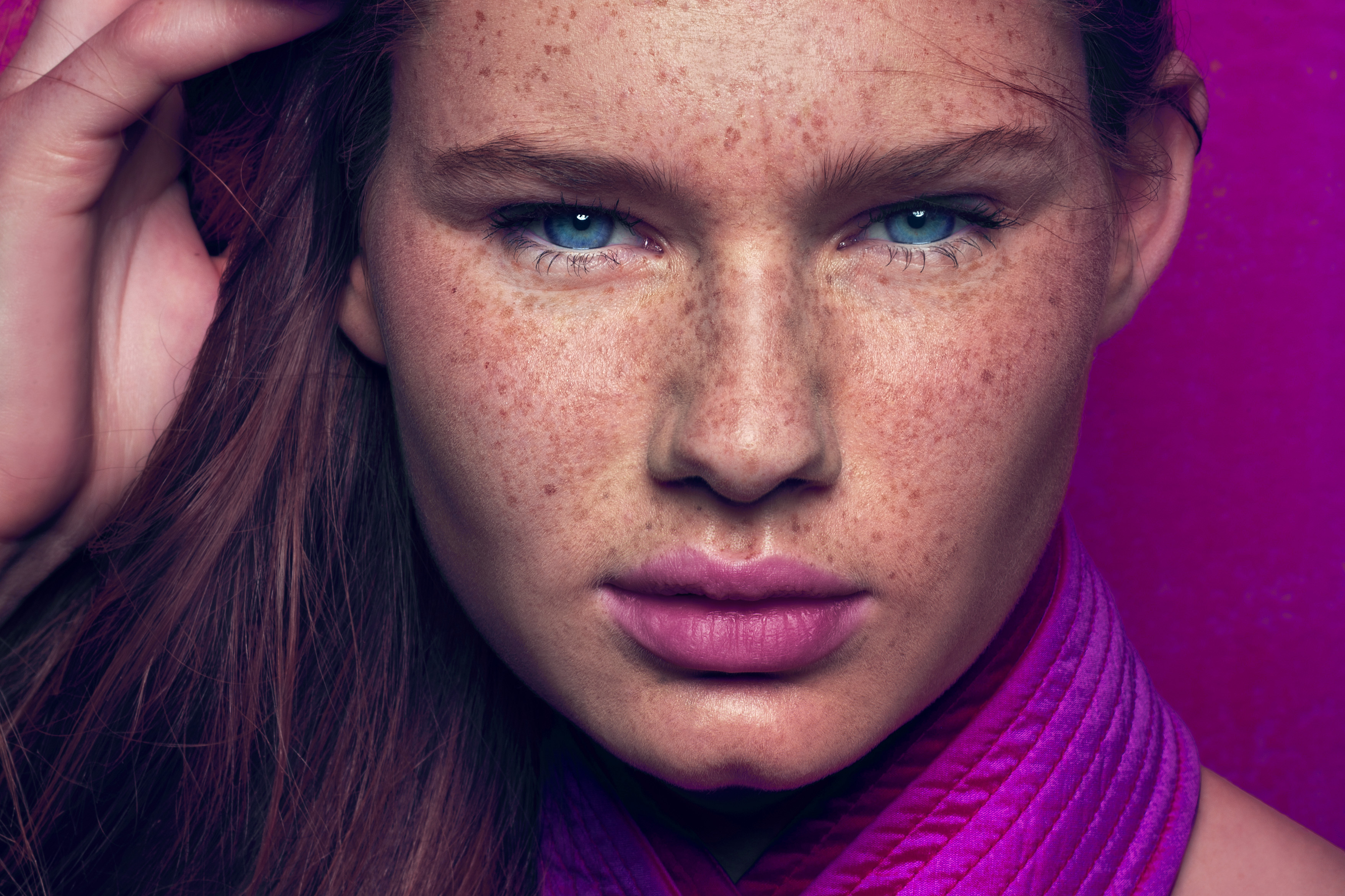 Close-up studio portrait of a red-haired woman with blue eyes and freckles against a vibrant magenta backdrop, wearing a pink top.