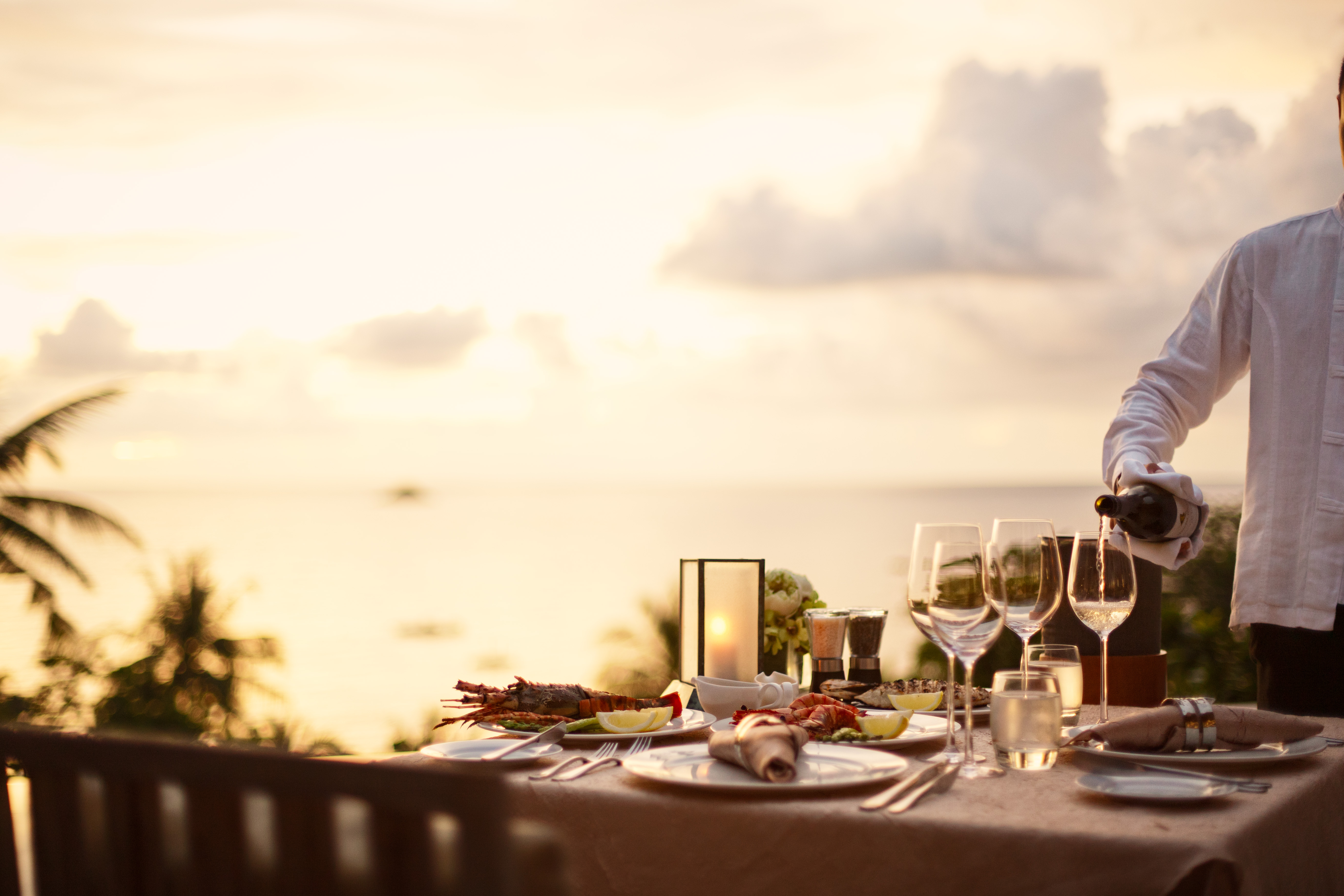 An outdoor dining table overlooking the ocean at sunset, set with wine glasses, plates, and food, being served by an adult male.