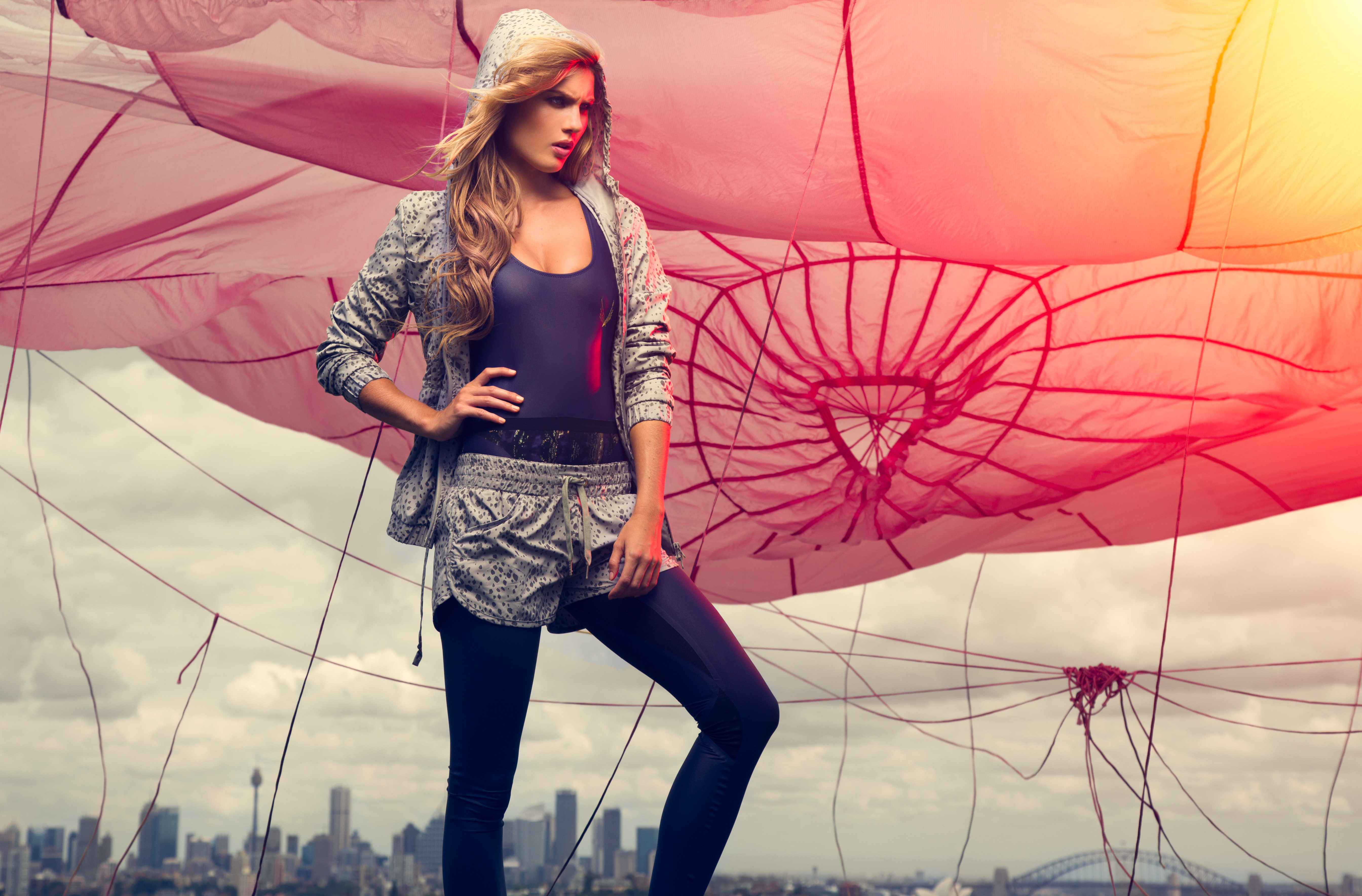 Adult woman in athletic clothing poses with hand on hip in front of city skyline and pink parachute-like structure.