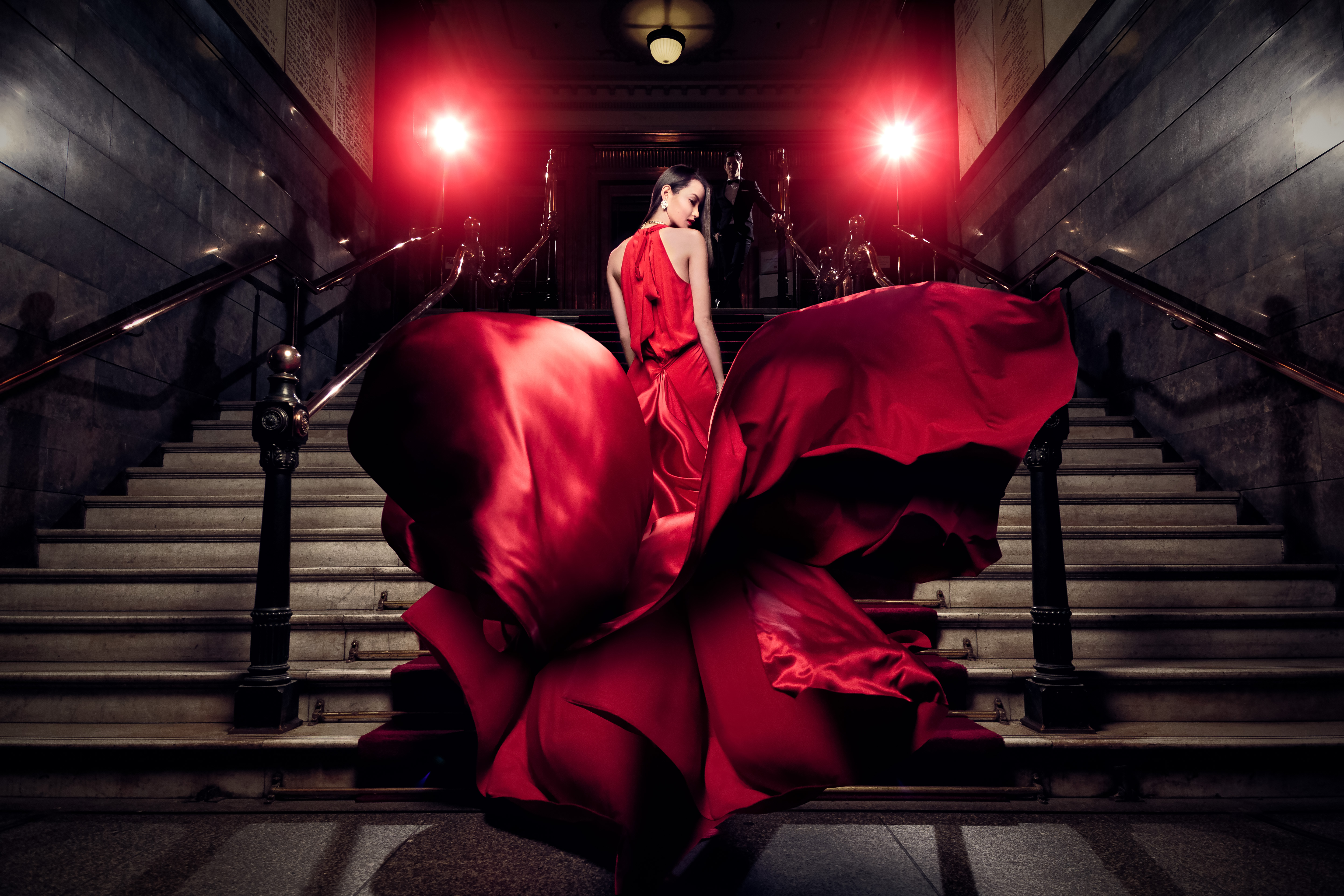 Woman in flowing red gown poses on marble staircase with red carpet and metal railing in dramatic, low-key lighting.