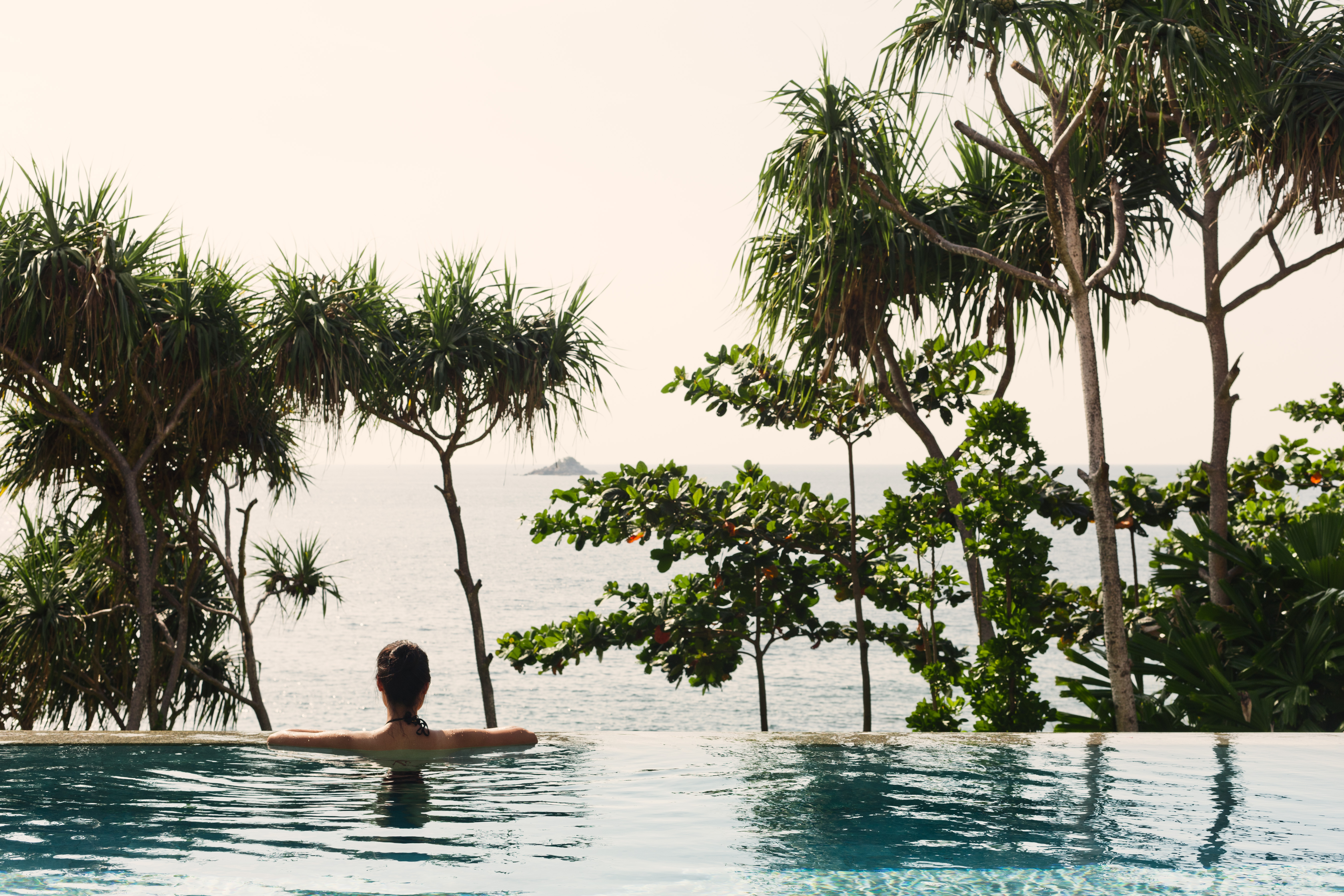 Woman relaxes in an infinity pool overlooking the ocean, framed by lush tropical trees, creating a serene vacation scene.