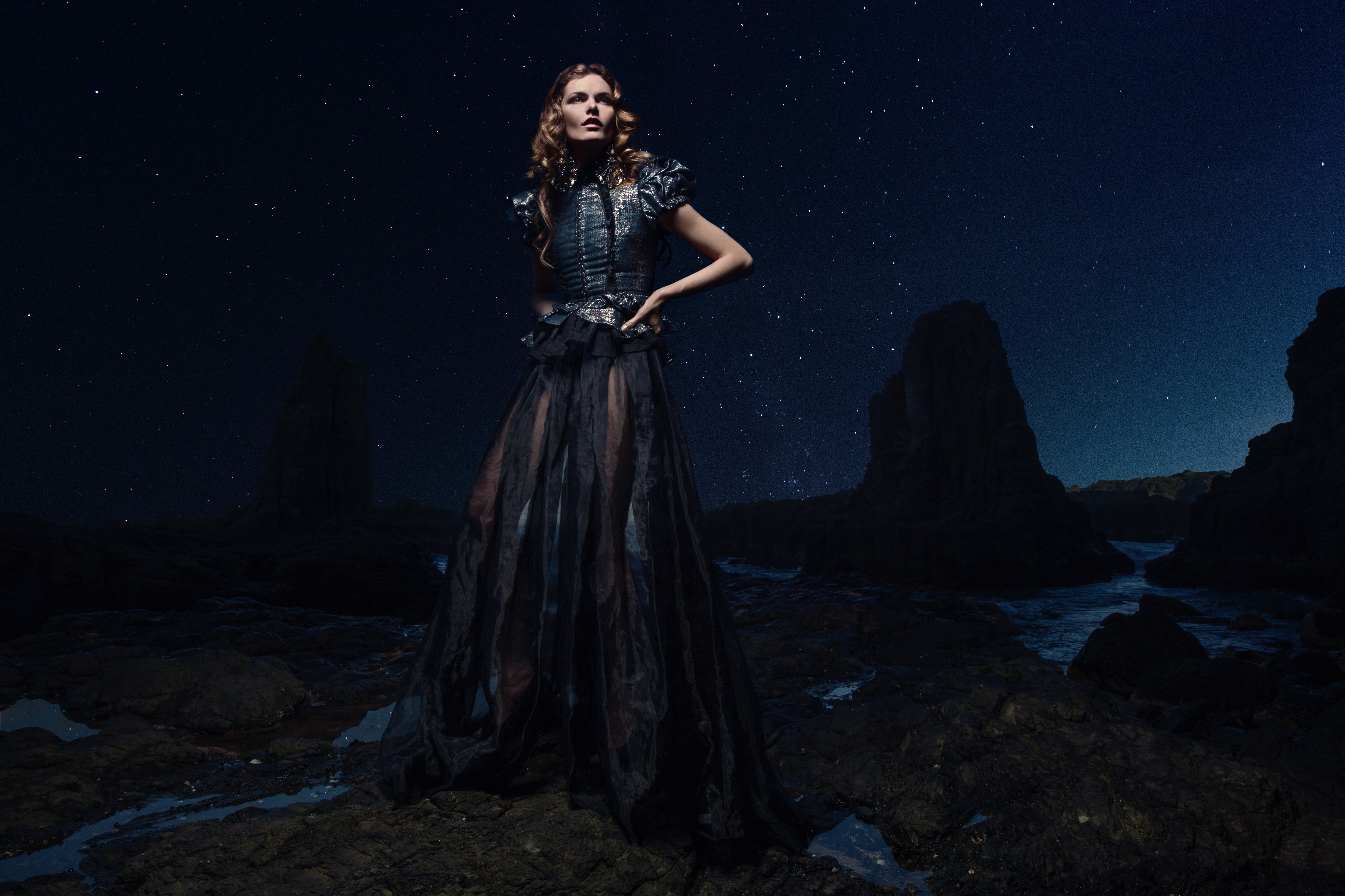 A model poses in a dark dress on a rocky shore under a starry sky, creating a dramatic fashion editorial with a blend of elegance and nature.