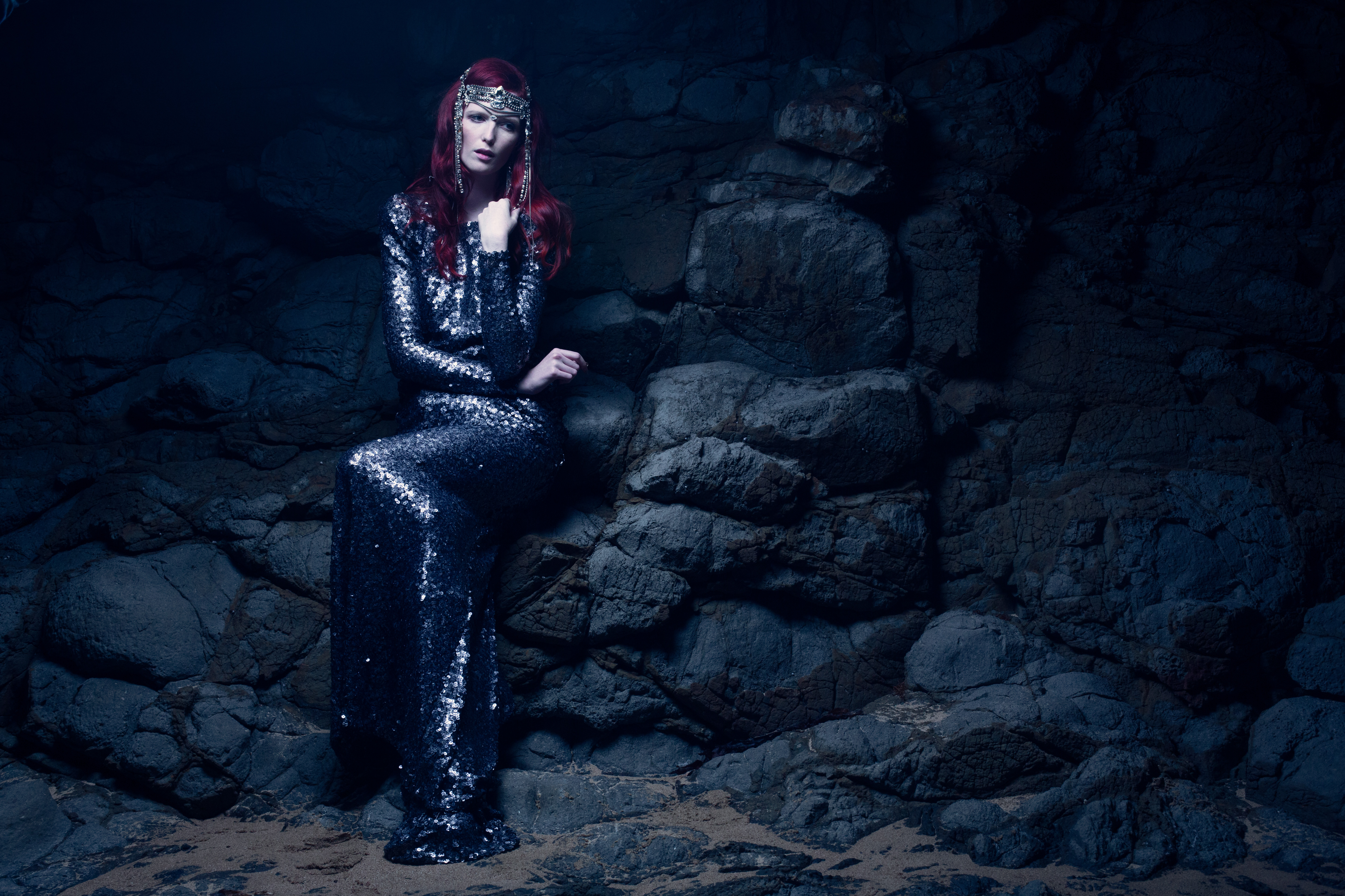 Woman in a sequin gown and headpiece sits on rocks in a dimly lit cave. The outfit sparkles under low lighting, creating a dramatic effect.