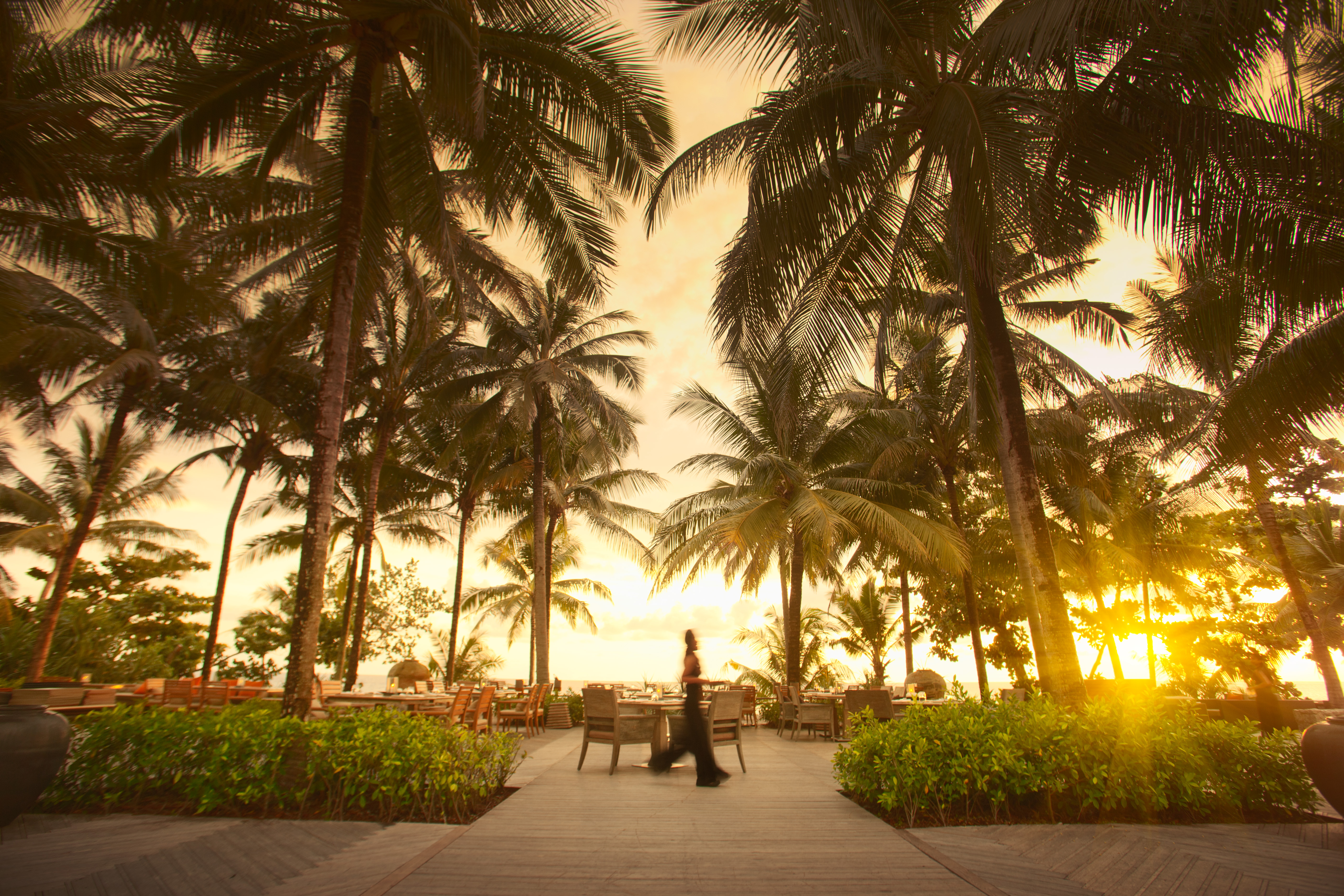 Woman walking along a wooden path lined with palm trees and greenery, leading to outdoor dining tables under a vibrant sunset sky.