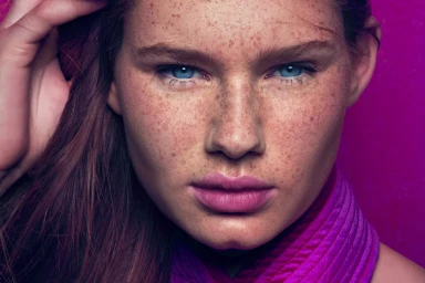 Close up portrait of a blue eyed woman with heavy freckles wearing purple clothing against a pink background