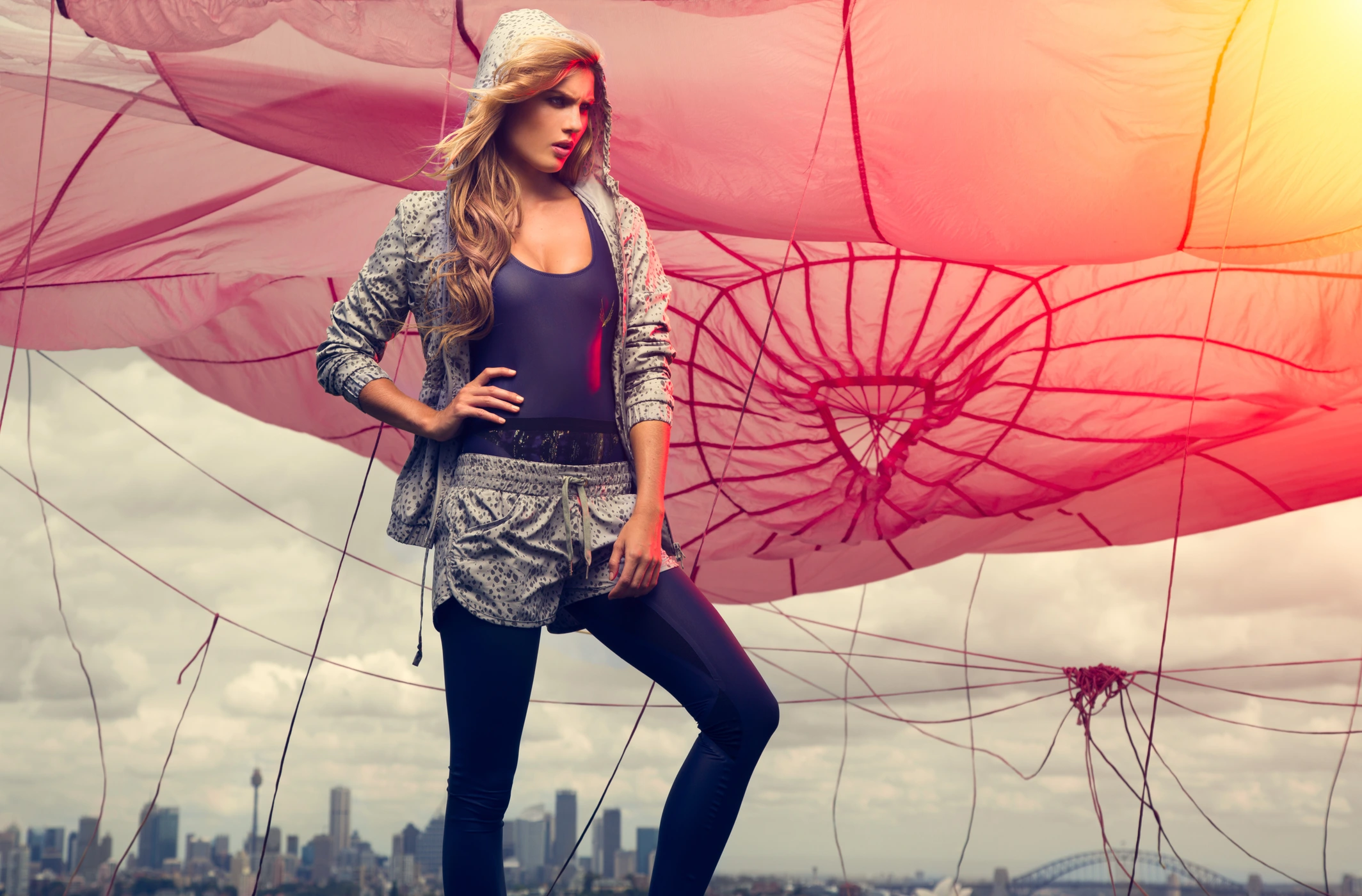 Model wearing patterned athletic hoodie and shorts standing against a large pink canopy with a city skyline below.