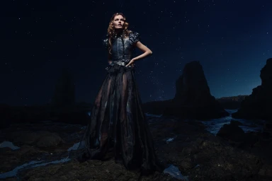 Model wearing a dark structured evening gown posing on rocky coast under a starry night sky, photographed by Andrew Maccoll.