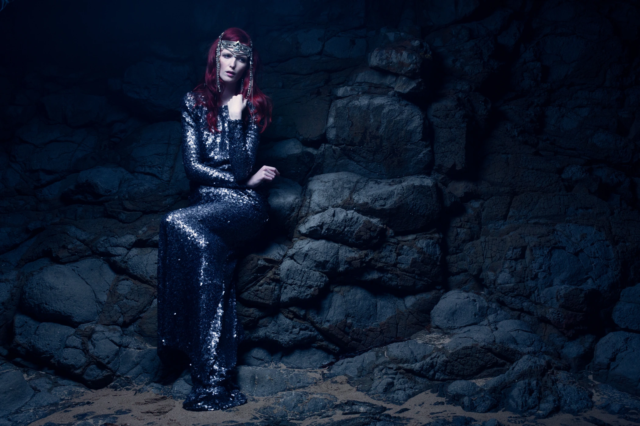 Woman with long red hair wearing a sparkly sequin dress seated against dark rocky background by Andrew Maccoll