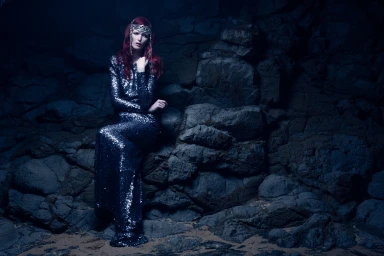 Woman with long red hair wearing a sparkly sequin dress seated against dark rocky background by Andrew Maccoll