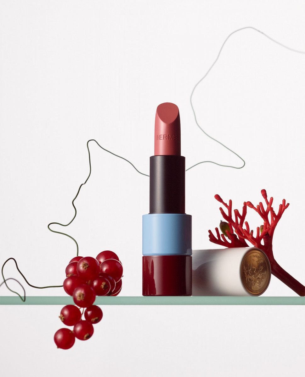 Close-up of a multi-colored lipstick tube with red berries and decorative twigs on a glass shelf against a white background.
