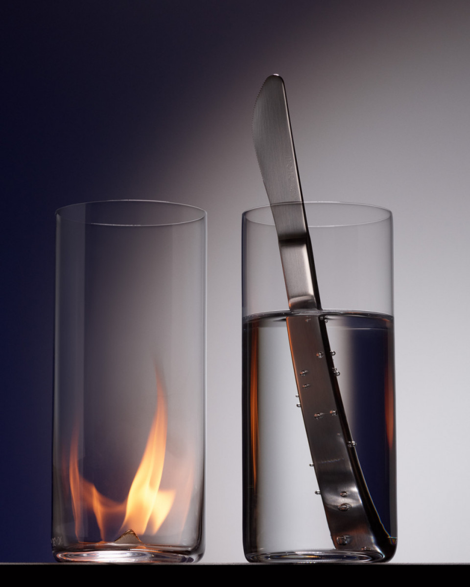 Still life of two clear glass cups: one containing a burning flame, and the other filled with water with a knife inside.