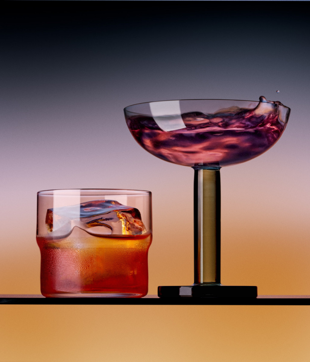 Two cocktails, one coupe glass with liquid splashing and one rocks glass with a large ice cube, stand on a reflective surface.