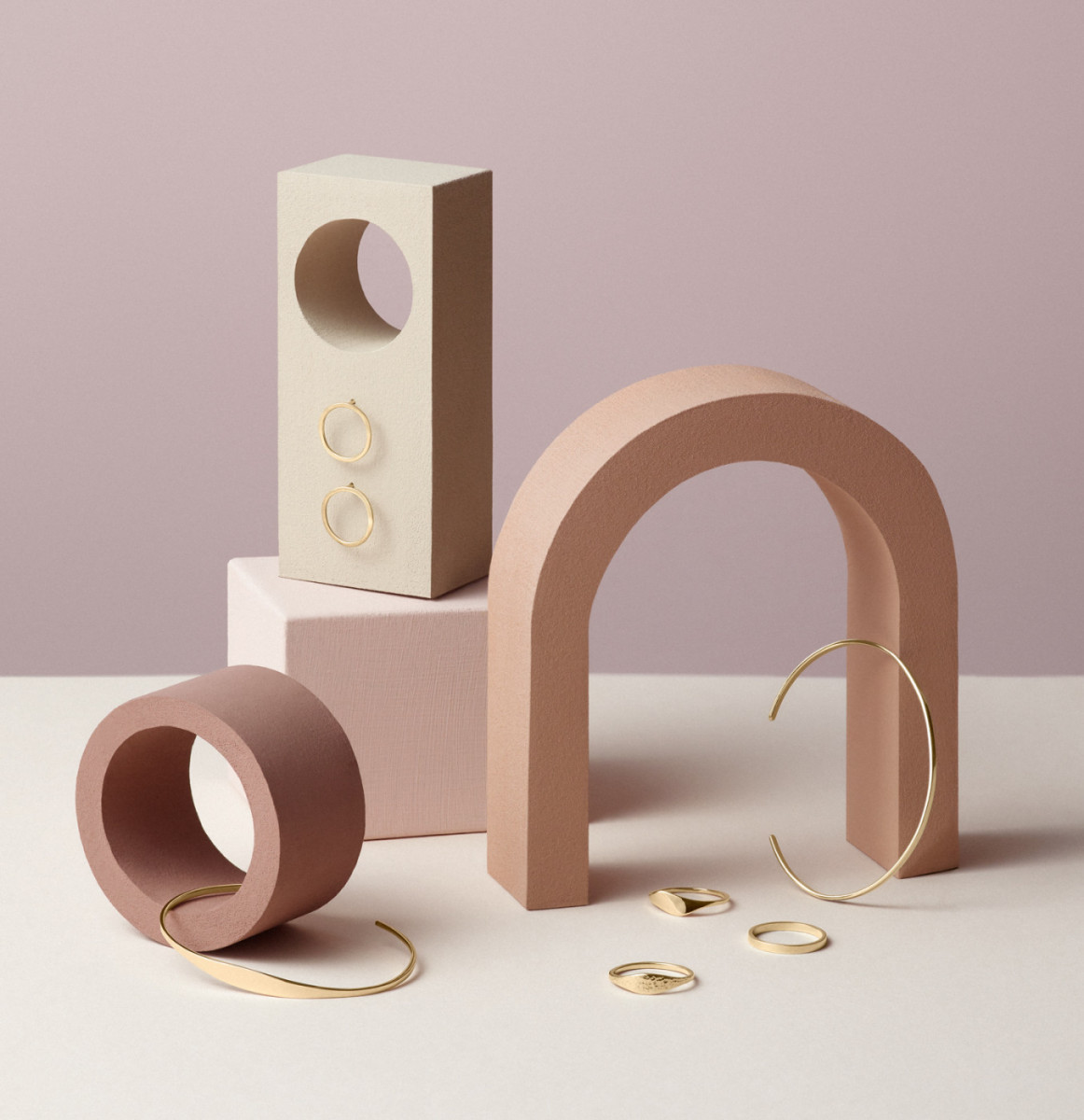 Still life of golden jewelry including rings, bracelets, and earrings, staged with geometric shapes in pastel colors.