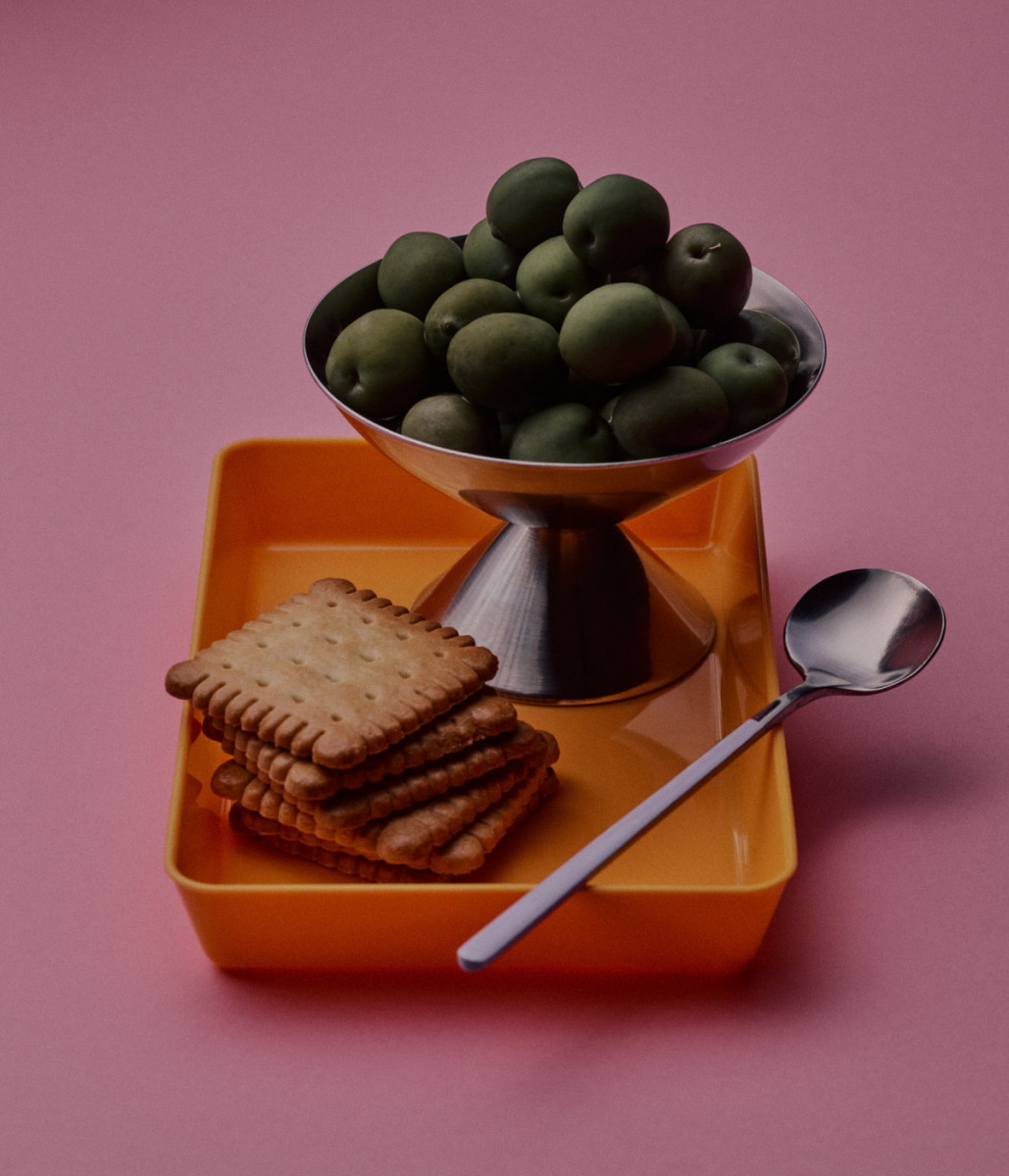 Elevated studio shot displays olives in a silver bowl, biscuits, and a spoon on an orange tray against a pink background, creating a vibrant food still life.