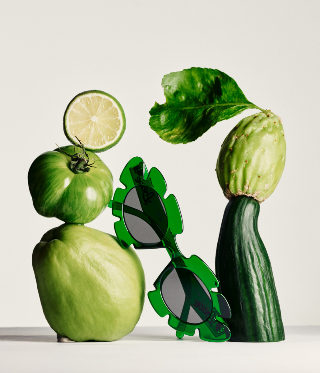 Still life arrangement featuring various green fruits and vegetables, including tomatoes, limes, and squash, alongside green sunglasses.