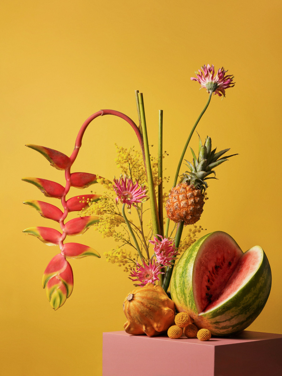 Still life composition with vibrant watermelon, pineapple, dragon fruit, lychee, and tropical flowers against a yellow backdrop.