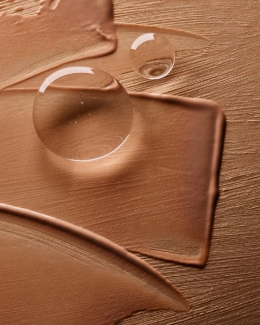 Close-up of a bronze-toned beauty product smear, featuring two clear water droplets for a modern, elegant aesthetic.