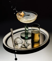 A cocktail in a coupe glass with a lemon twist, a highball glass, ice cubes, and tongs on a metallic oval tray against a dark background.