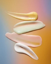 Four colorful cosmetic cream smears displayed vertically on a gradient pastel background