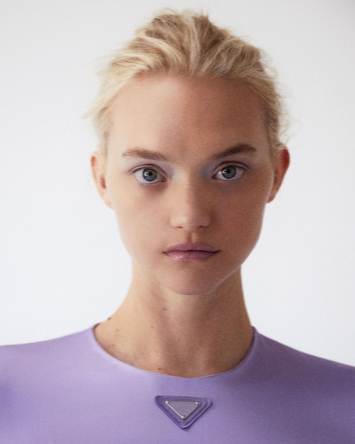 Close-up portrait of a young adult woman with blonde hair and blue eyes, wearing a lilac top, against a light grey background.