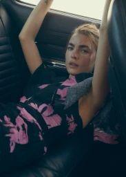 Model with arms raised lying on the back seat of a car wearing a dark dress with pink floral print.