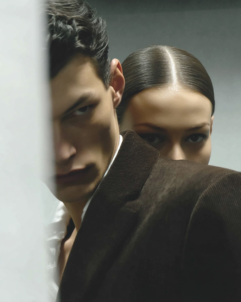 Portrait of a stylish couple: a man in a blazer and a woman with slicked-back hair, posing against a neutral backdrop.