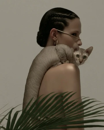 Woman in profile holding a hairless cat over her shoulder in a studio with neutral tones and some palm fronds in the foreground.