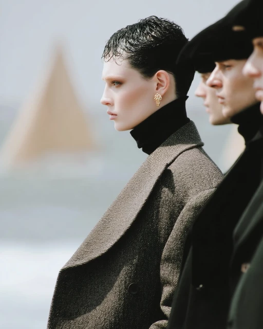 A diverse group of fashion models standing in a line in an outdoor setting, wearing black turtlenecks and stylish coats for an editorial shoot.