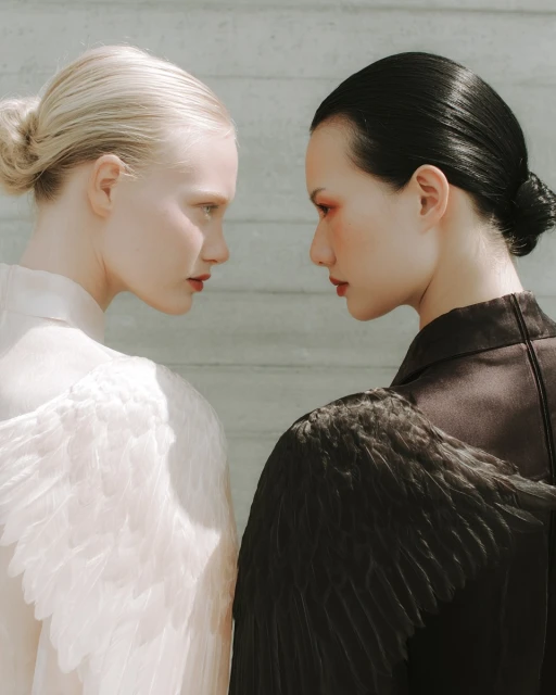 Two adult women pose with black and white feathered wings against a neutral background, showcasing contrasting fashion and beauty styles.