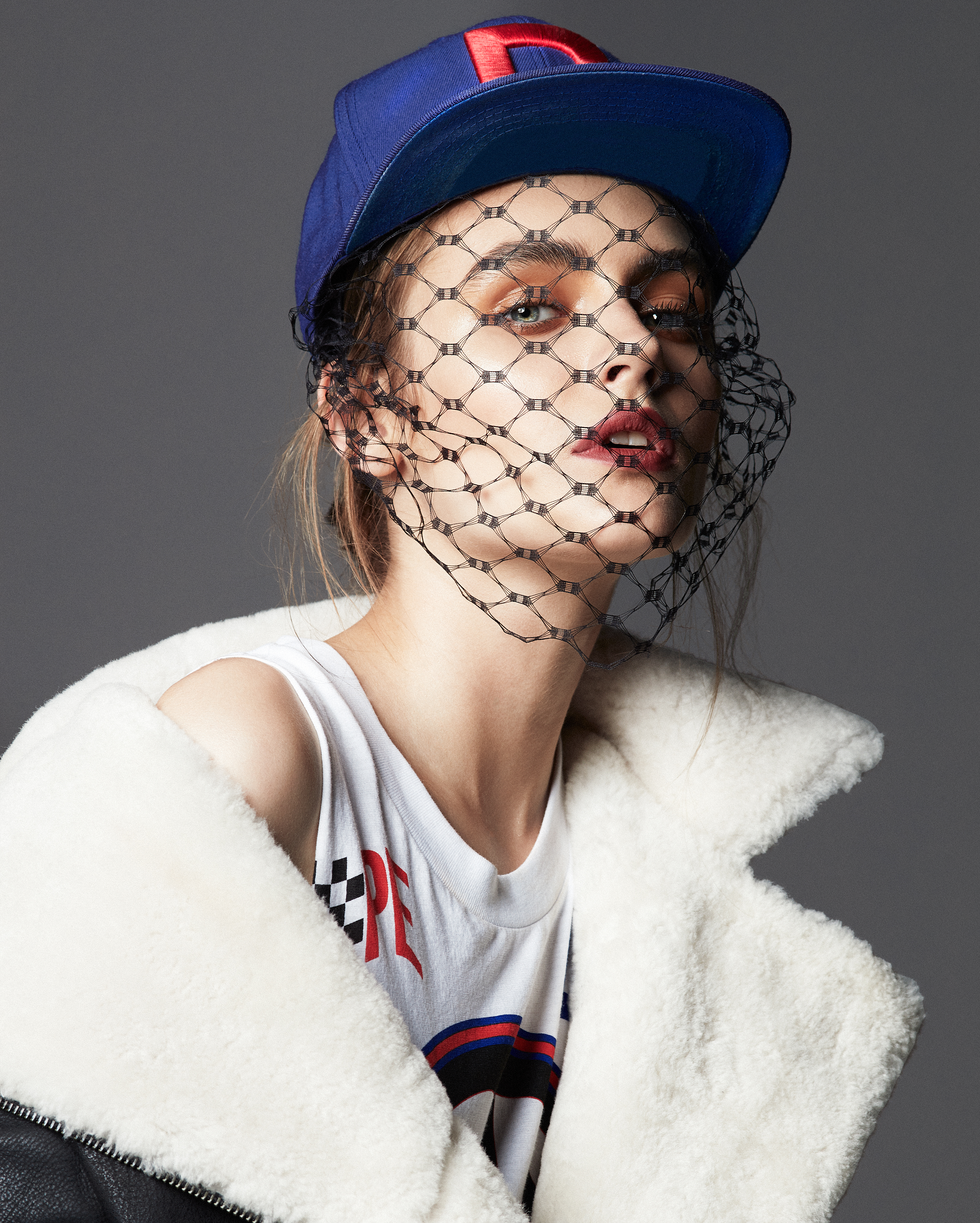 Woman wearing a baseball cap and black veil, posed in a studio with a sheepskin jacket. Fashion style with cool mood.