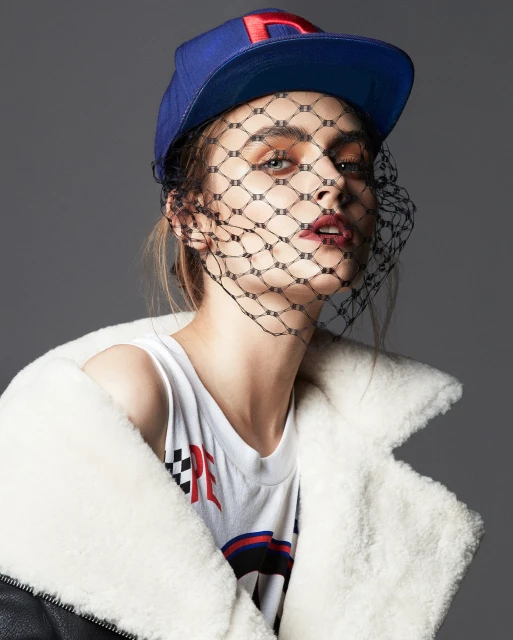 Woman wearing a baseball cap and black veil, posed in a studio with a sheepskin jacket. Fashion style with cool mood.