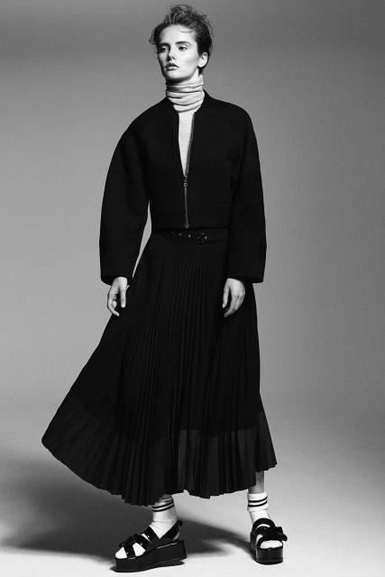 Monochrome fashion shot of a woman wearing a black jacket, pleated skirt, socks, and sandals against a neutral studio backdrop.