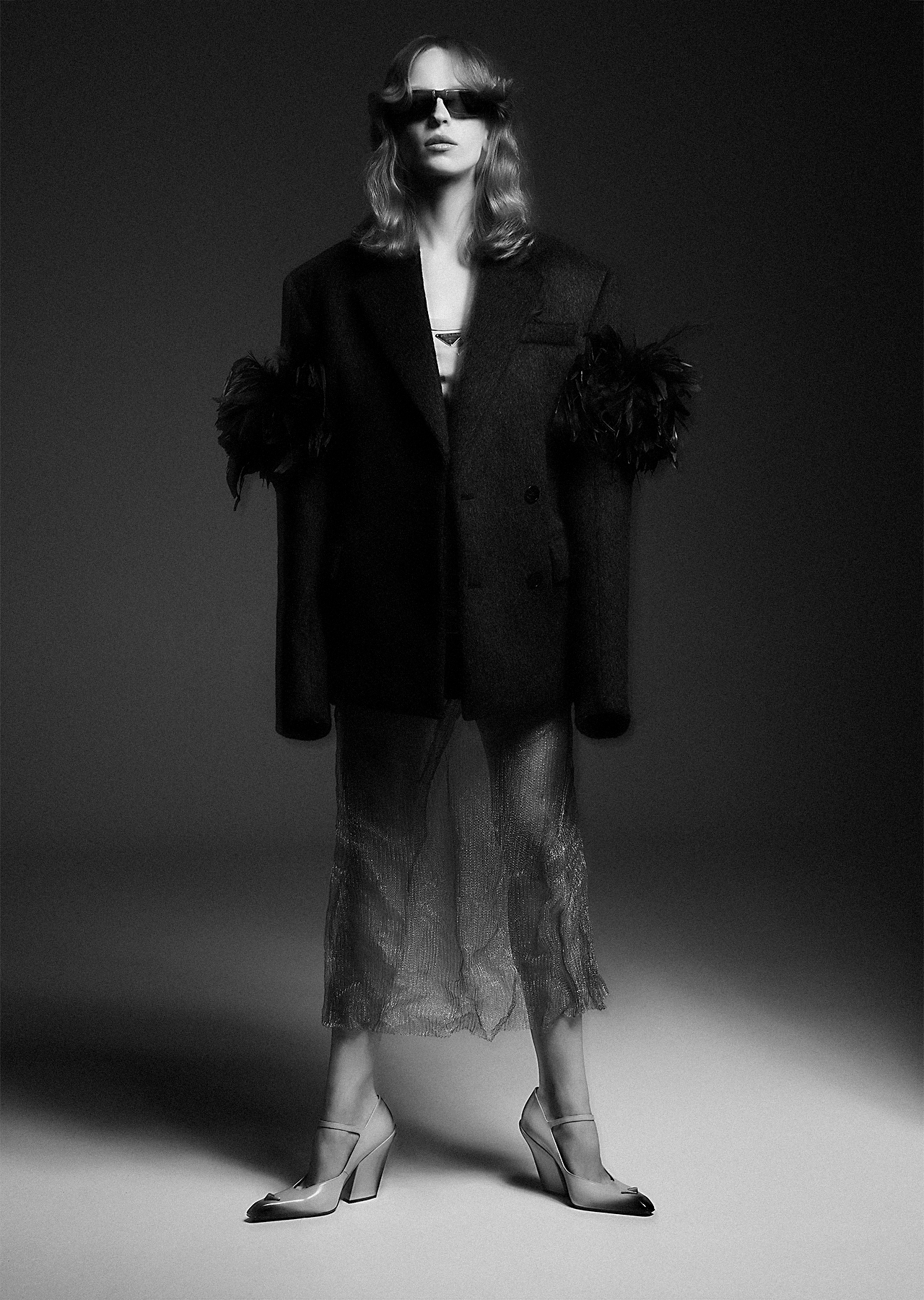 Monochrome fashion shot of a woman in an oversized black blazer with feathered sleeves and high heels posing in a photo studio.