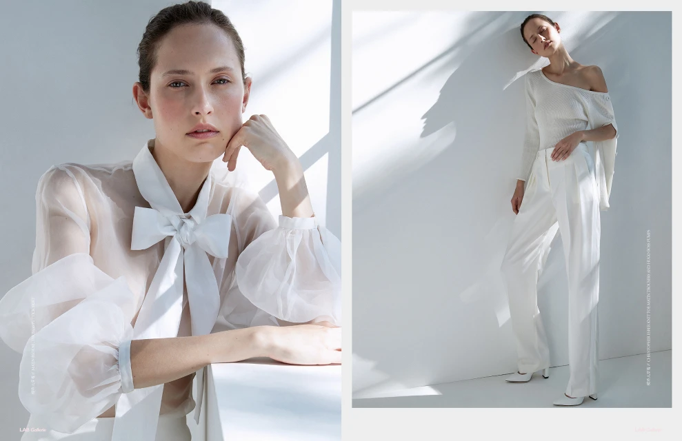 Fashion shot of a woman in a white top with a large bow and white trousers leaning against a white wall in a light, bright studio.