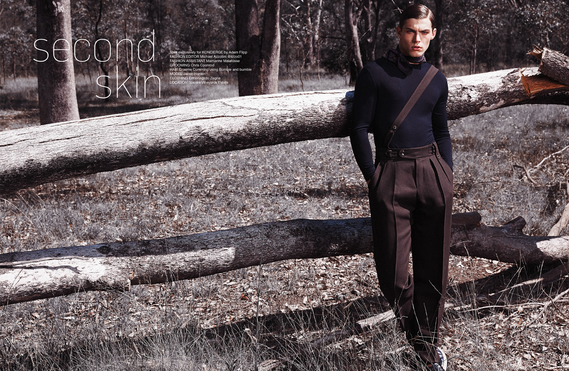 Fashion editorial shot outdoors with a young male model in dark, formalwear, leaning against a fallen tree trunk in a forest setting.