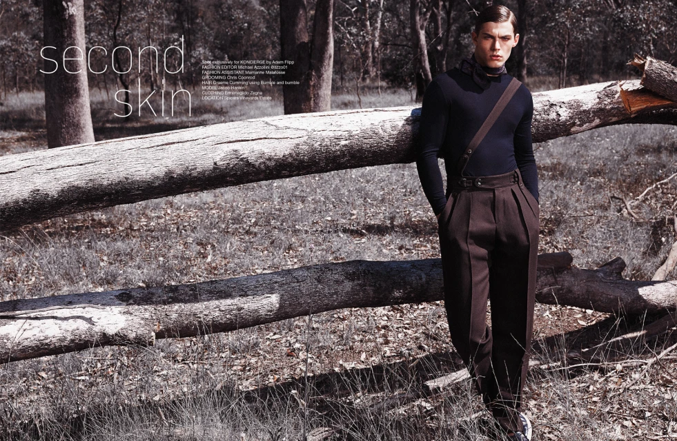 Fashion editorial shot outdoors with a young male model in dark, formalwear, leaning against a fallen tree trunk in a forest setting.