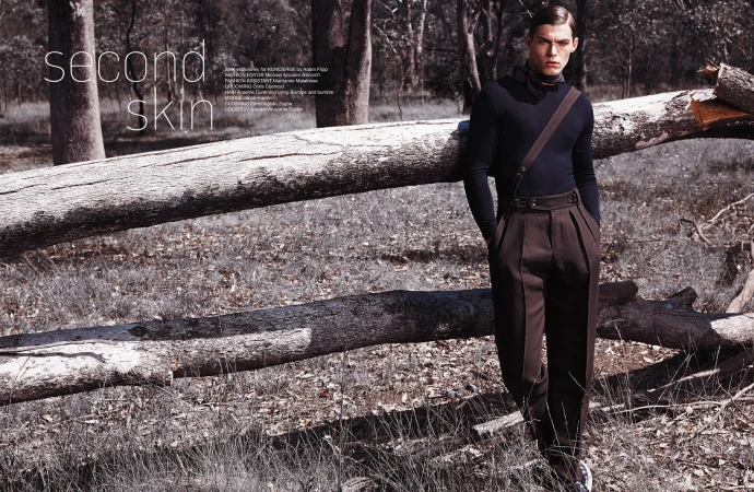 Fashion editorial shot outdoors with a young male model in dark, formalwear, leaning against a fallen tree trunk in a forest setting.